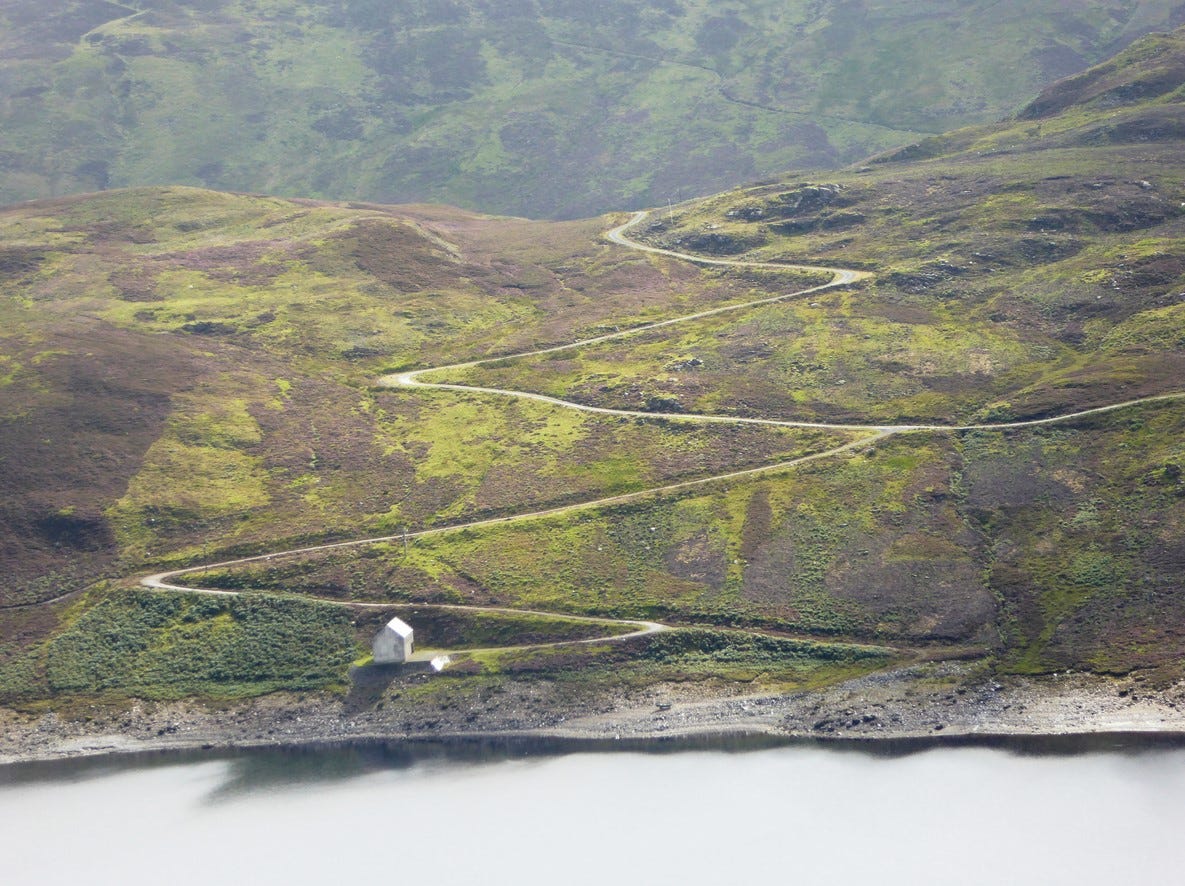 an image of a hill with a zigzag path carved into it, meeting a lake and a small building at the bottom.