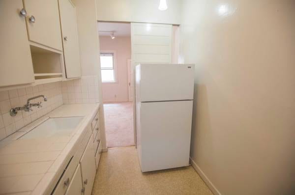 A very small kitchen with a small white refrigerator blocking half of the door to another room.