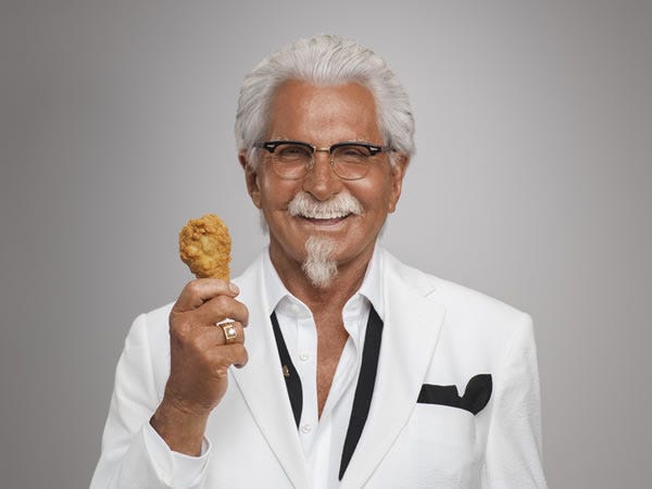 Colonel Sanders smiling and holding up a drumstick, his shirt collar unbuttoned and tie loose.