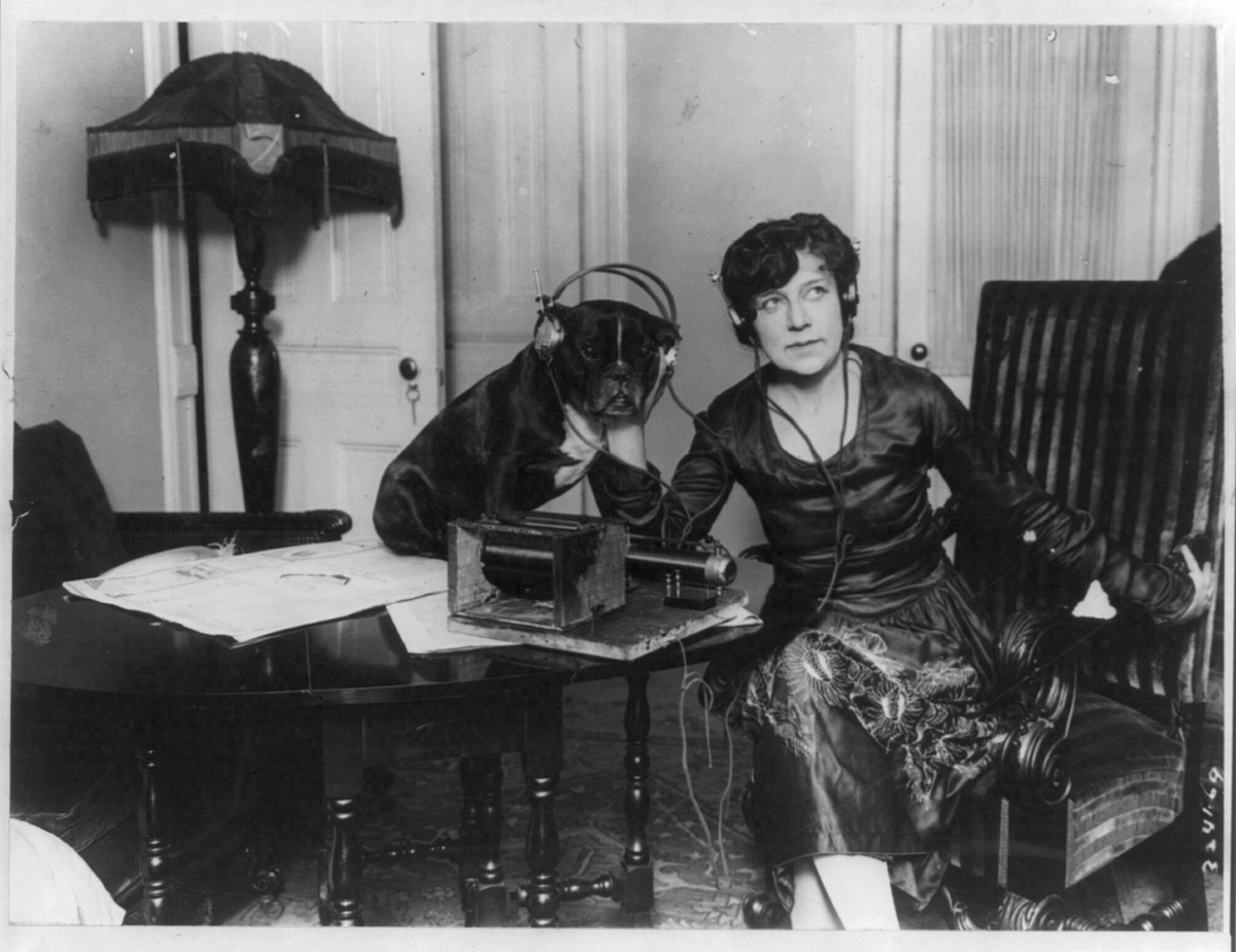 Black and white photo of Mme. Asta Souvrina, “the Bernhardt of Russia,” sitting beside a dog named Buster. Both are wearing headphones, listening to a radiophone device on a table in front of them. Taken around 1922 by Underwood & Underwood.