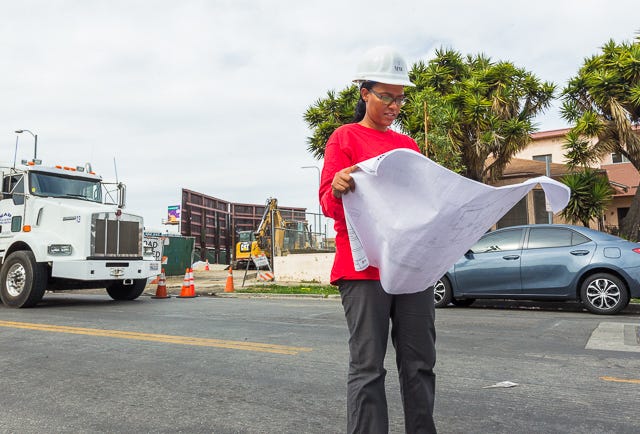 Mel Wyatt in a red longsleeve shirt and white hard hat, looks at plans at a Los Angeles project site.