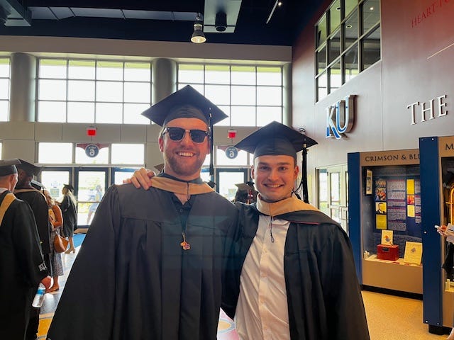 Two men pose for a photo while wearing graduation regalia.