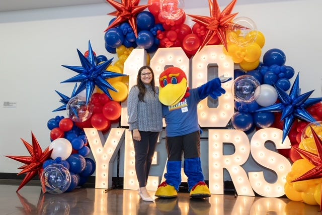 A girl poses with Baby Jay in front of light up letters that read "100 years" and surrounded by red, blue and yellow balloons.