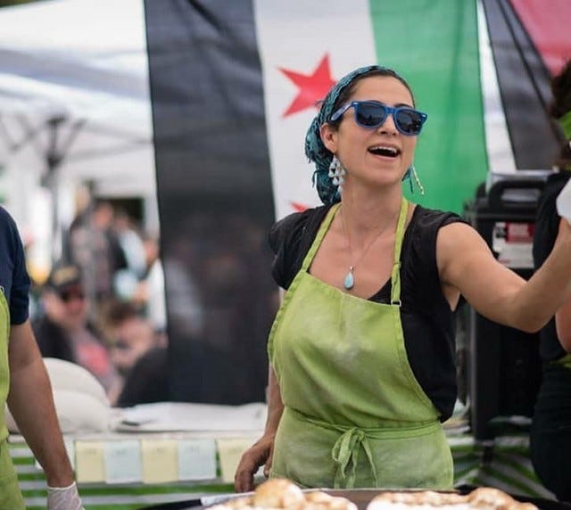 A person wearing a green apron and dark shades, hair pulled back, speaking outside.