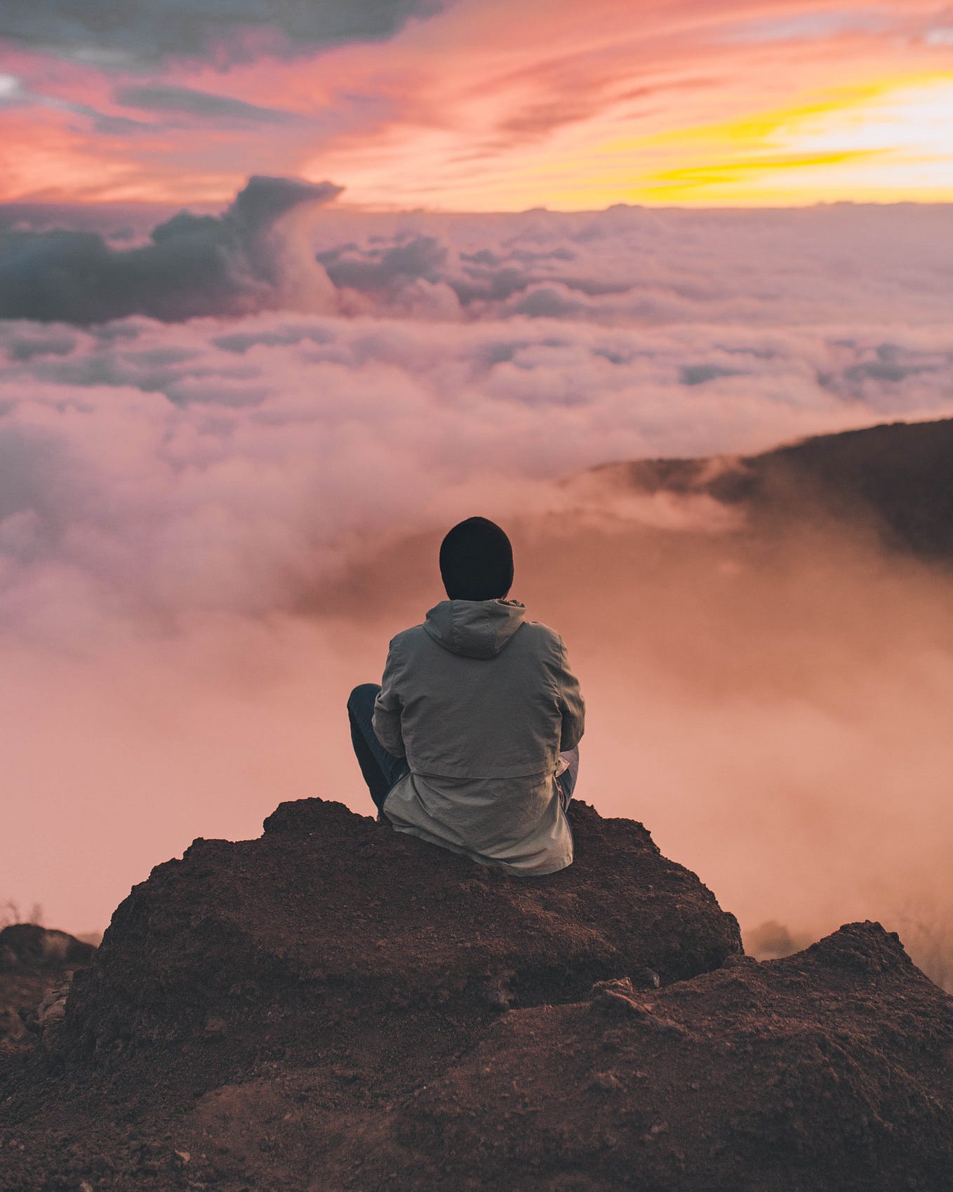 A man sitting cross-legged on a mountain top looking at the pink colored cloud canopy underneath his eyesight