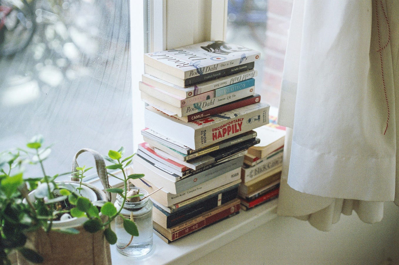 A stack of books sitting on a window ledge with the light streaming in. There is a flowing white curtain and a house plant, one on either side of the stack of books.
