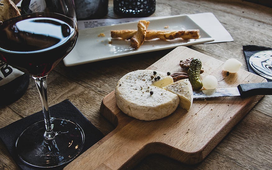 Dark red wine in a tall wine flute on a black napkin next to a wooden cutting board with a cheese wheel, a pie slice cut out, adjacent to a knive and bits of herb and cheese. Behind the cutting board is a ceramic tray with a twisted bread stick on it.