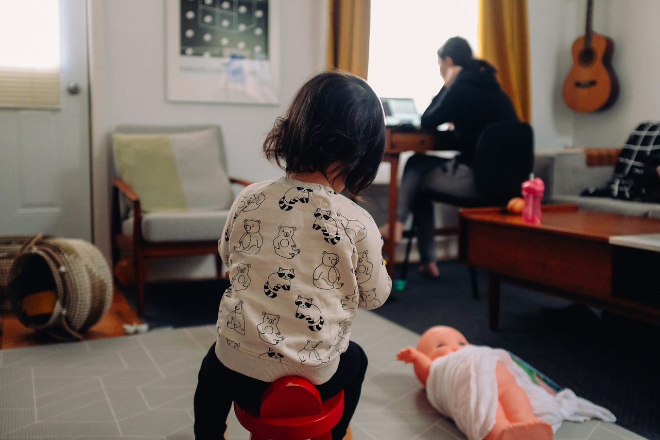A little girl sits on a stool, facing her mother who is working at her desk, back turned to her.