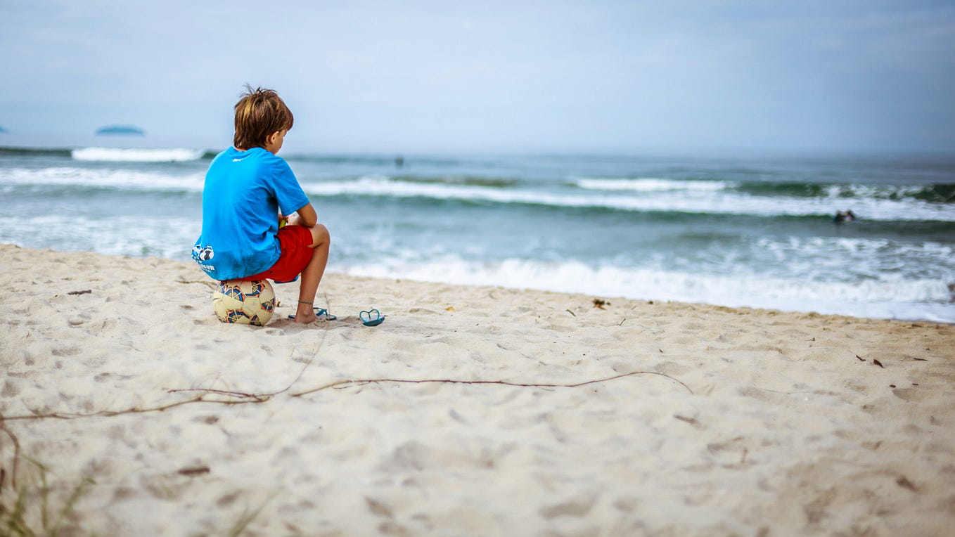 A little boy sits along staring at the waves along the shore.