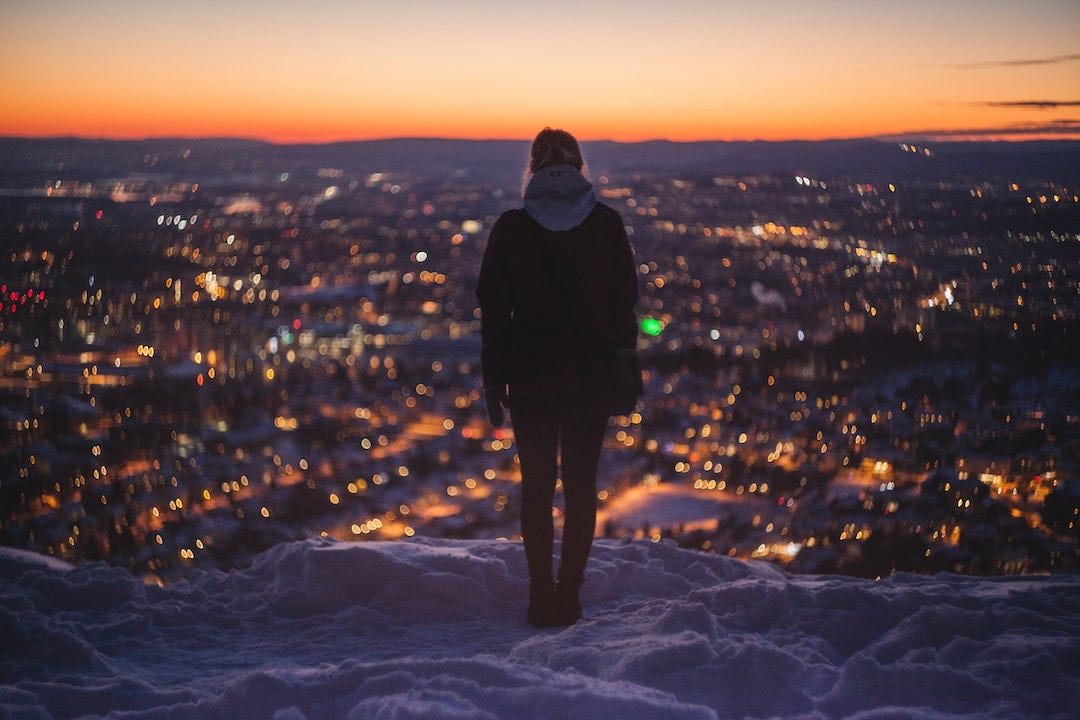 A woman stands near the edge of a cliff overlooking a city.