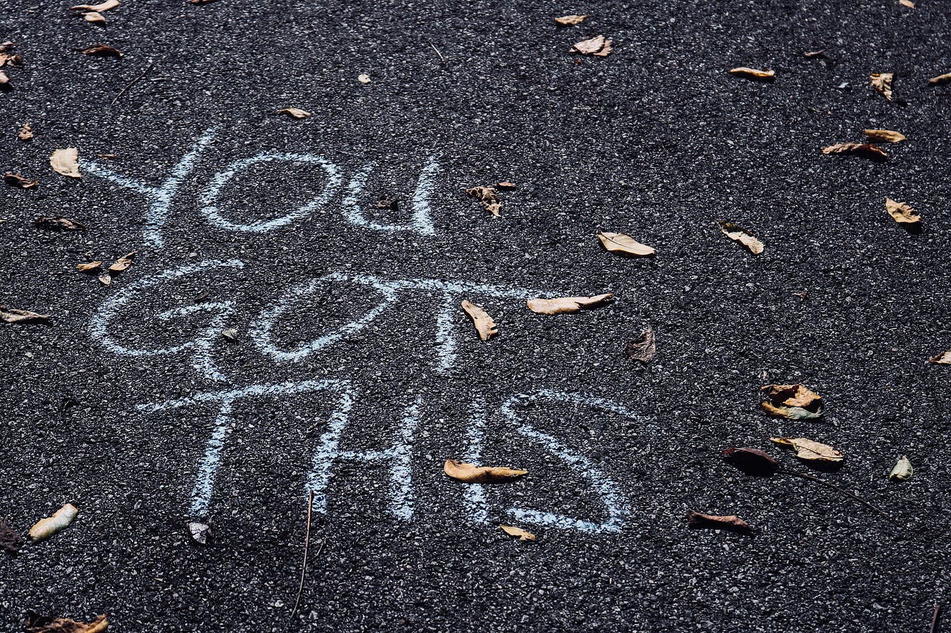 The words “YOU GOT THIS” are written in chalk on an asphalt surface. A few dried leaves are scattered on the ground around the writing.