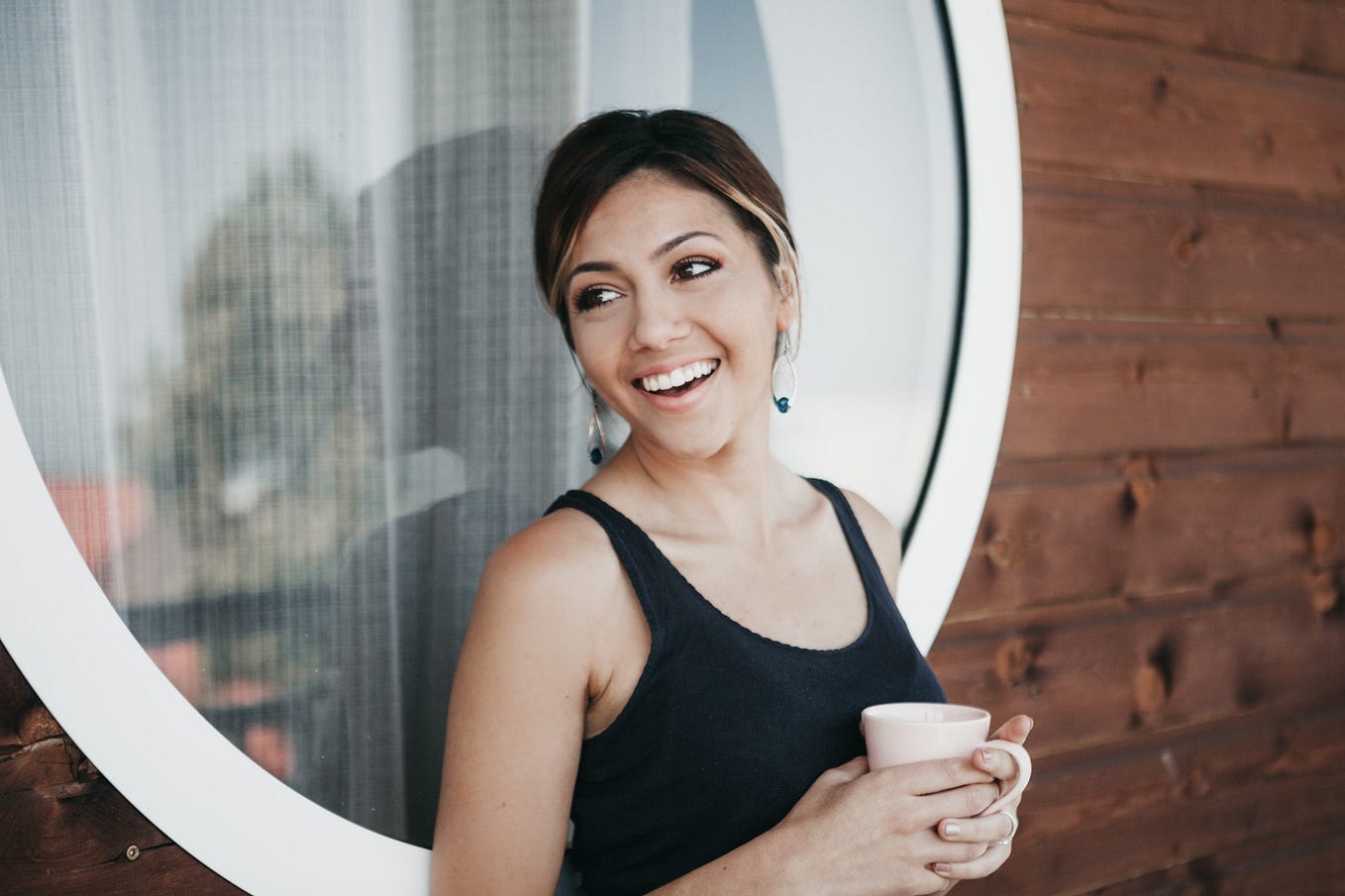 Woman leaning against a window with a mug in her hand and a smile on her face.