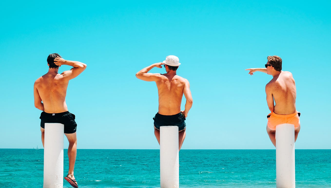 Three guys in swimming trunks sitting on white plinths, looking out onto an aqua sea on a sunny day.