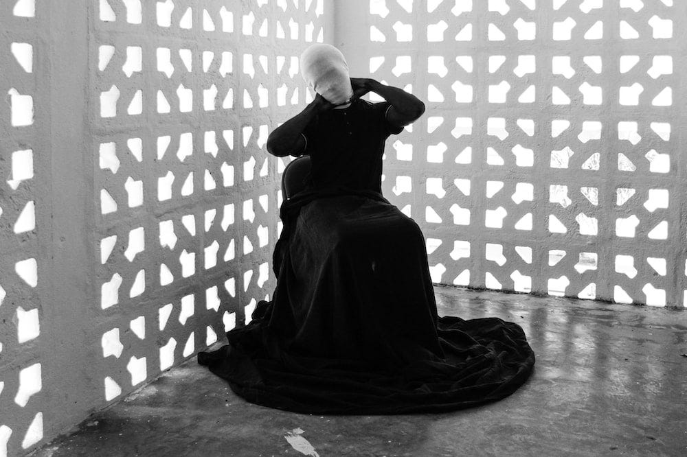 A black and white photo of a woman with a long black dress sitting in a chair. A white cloth mask is covering her face she’s sitting in front of a white fence.