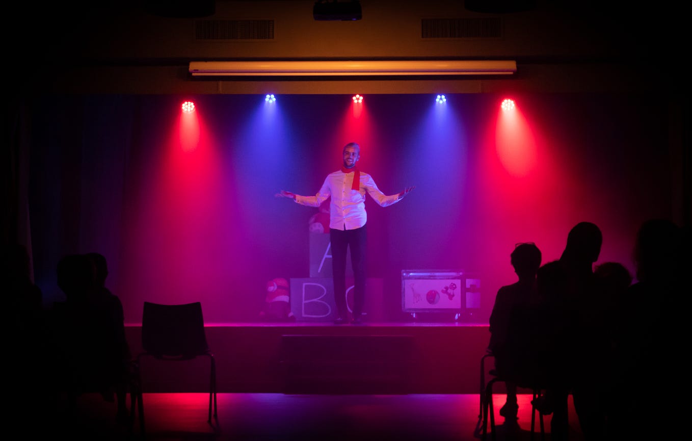 A white man with a beard wearing a white collared shirt, red scarf, and dark pants stands on a stage backlit by alternating blue and red lights in an otherwise dark room. He has his arms slightly outstretched. The dark silhouettes of a few people in the audience can be seen.