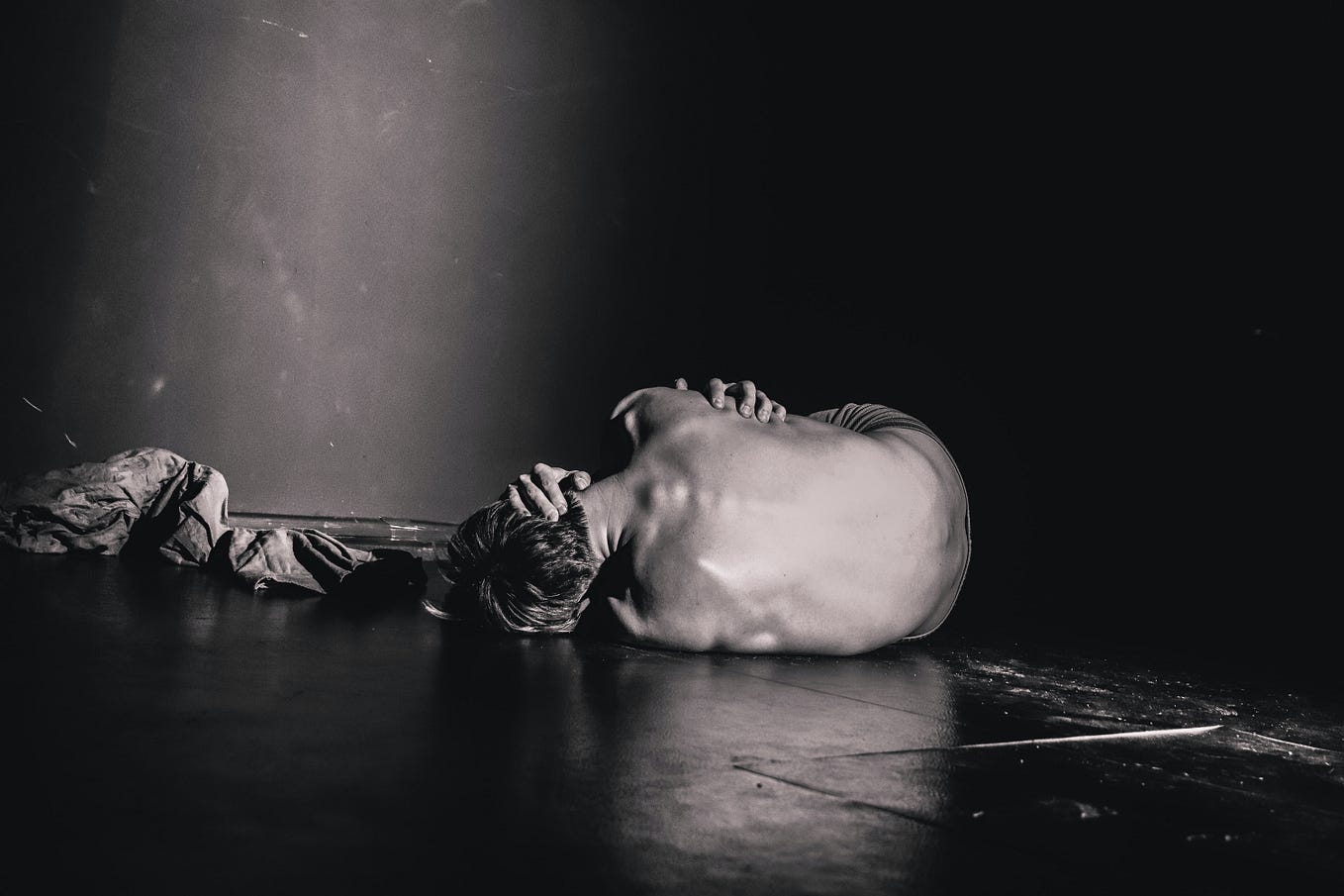 A black and white photo of a person lying on their left side in the fetal position hugging themselves for comfort on a wooden floor.
