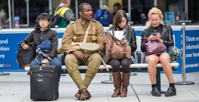Rail travellers sit on a bench with We’re Here Because We’re Here participant dressed as WW1 soldier.