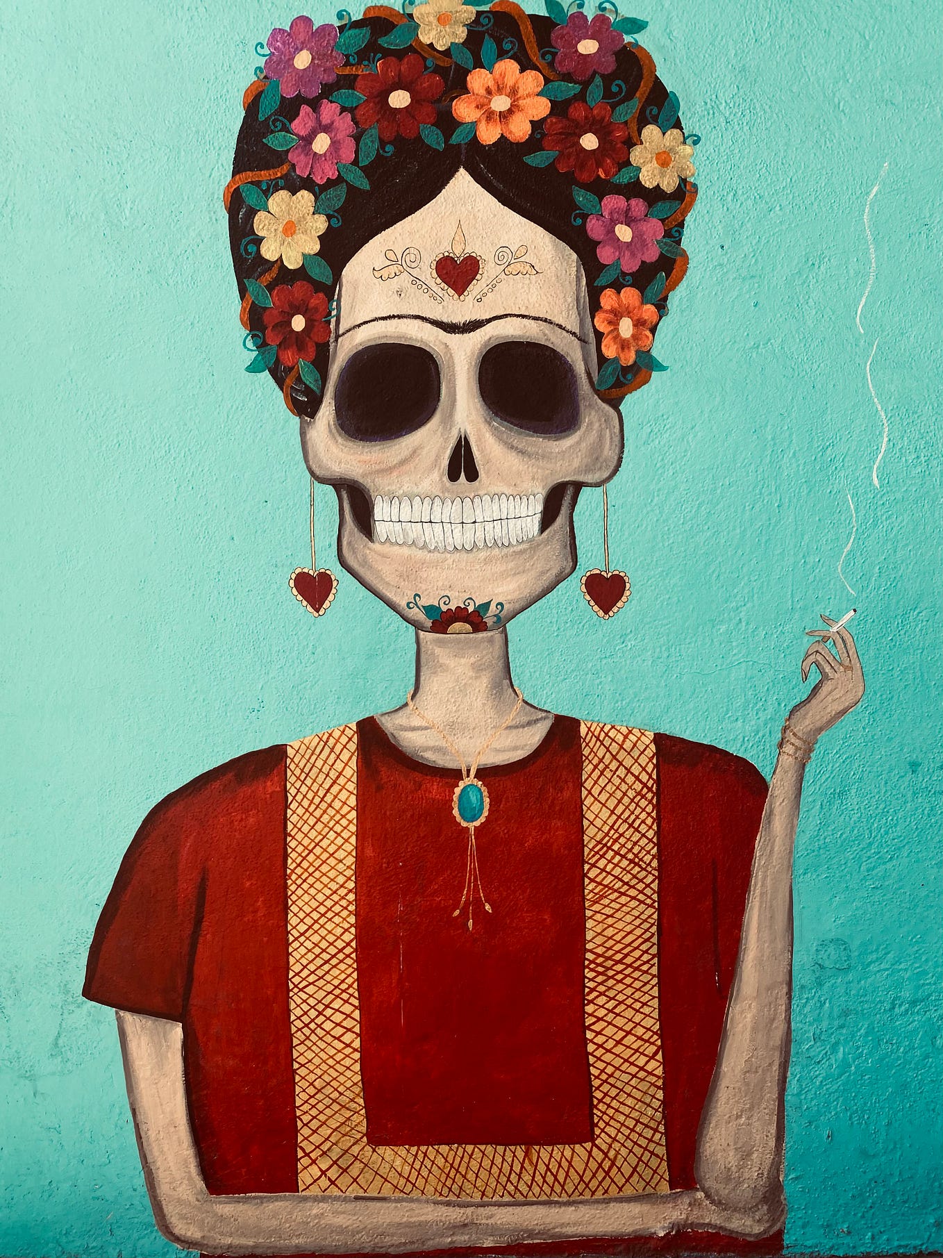 A mural on a blue wall. The mural depicts a sugar skull — a female-appearing skeleton covered in flowers and hearts. She is smoking a cigarette and grinning. Her unibrow suggests that she is Frida Khalo, or at least an homage to her.