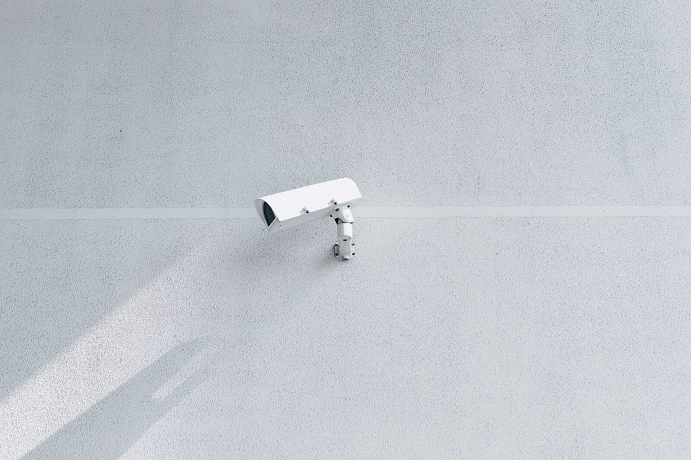 A photograph of a CCTV surveillance camera attached to a blank wall and its shadow