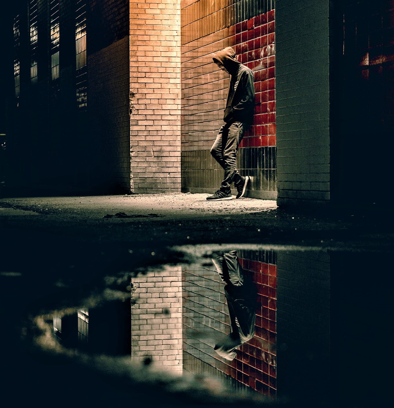young man wearing hooded sweatshirt leaning against brick wall at night with reflection in puddle