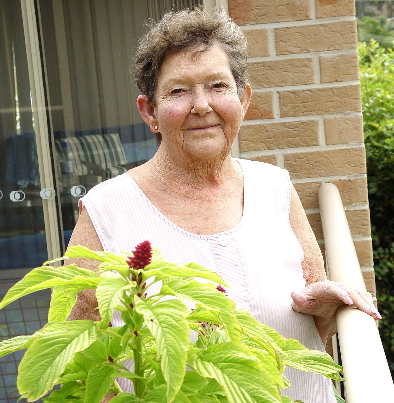 Elderly woman looking at the camera with a gentle smile on her face. One hand rests on a balcony railing and a plant with a red flower is in the forefront.