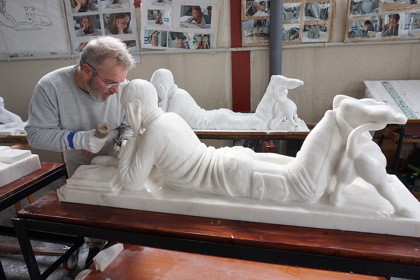 A sculptor chiseling away a block of marble to reveal a statue of a man, laying on his stomach with his head in his hands.