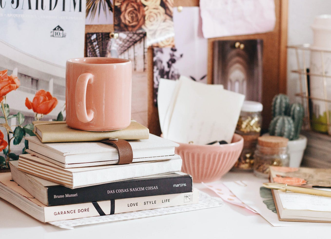 A stack of books and journals sits on a white desk with a pink mug on top of the stack. The desk is covered in a variety of items, including a cactus plant, small pink bowl, and papers. There is a corkboard in the background mostly obscured by photos of flowers and architecture pinned to it.