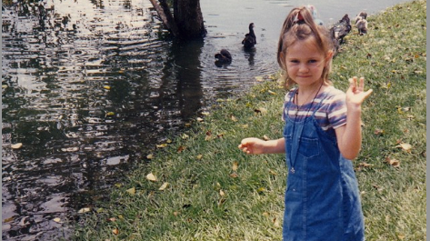 A young girl waves alongside the edge of a lake, with ducks in the distance.