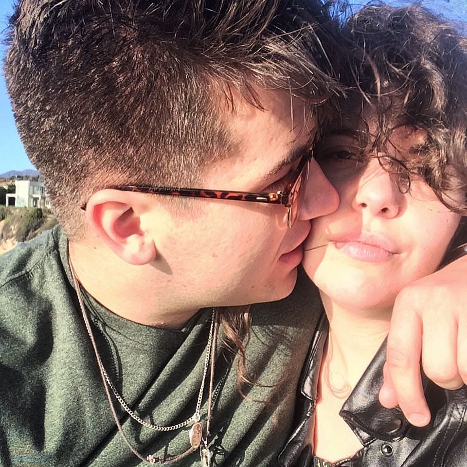 Man wearing sunglasses kissing woman’s cheek with arm around her