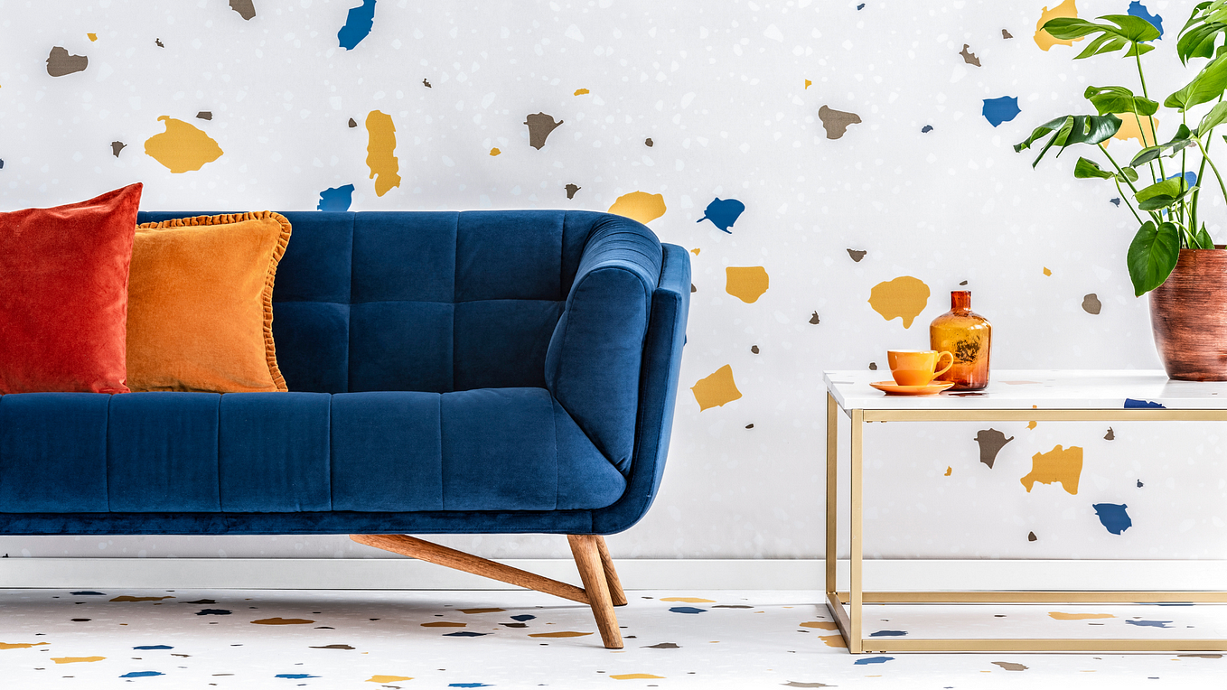 Royal blue MidCentury Modern couch with peg les sitting in front of a confetti colored wall.