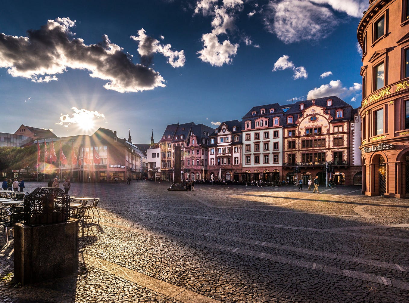 A photo of a plaza with cobblestone pavement and old-looking buildings with southern German architecture. The sun is low on the horizon with clearly distinct rays beaming across the square, creating long shadows on the pavement.