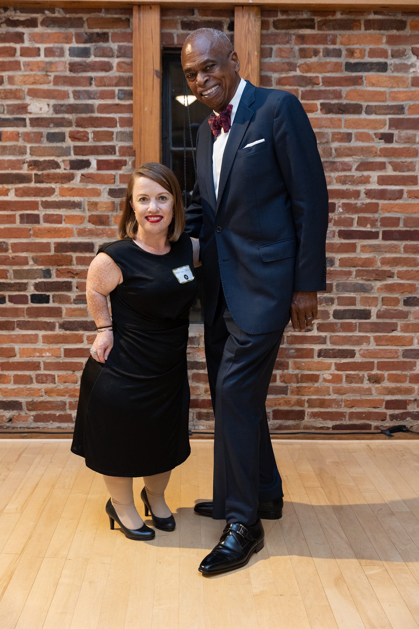 A white redheaded dwarf woman in a black dress standing next to a tall African-American man wearing a blue suit.