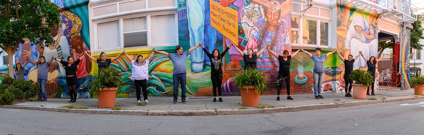 Group of performers on a sidewalk in front of a street mural holding up linked hands
