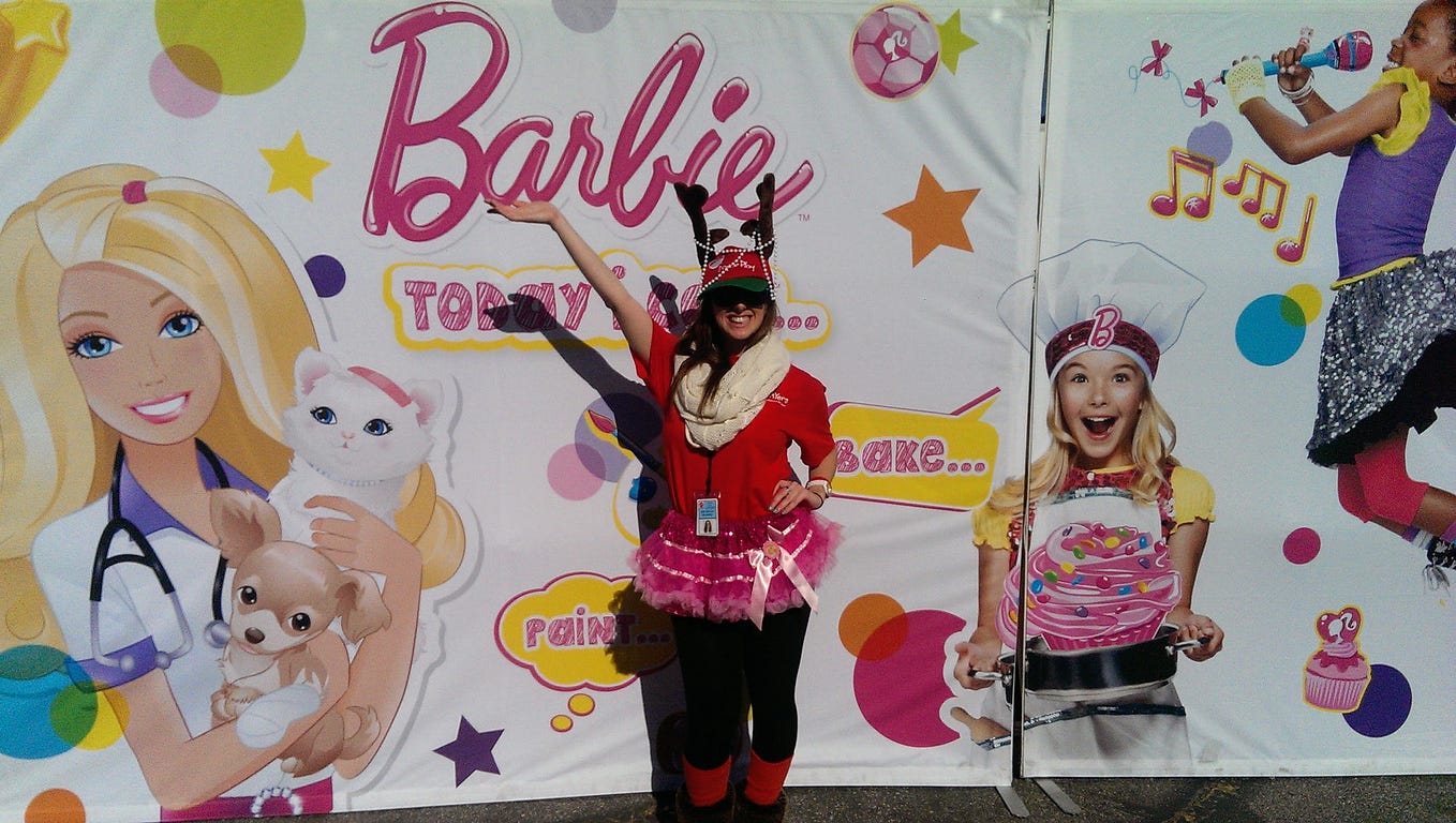 A girl in a red shirt with a reindeer hat, scarf, and tutu stands in front of a Barbie poster.