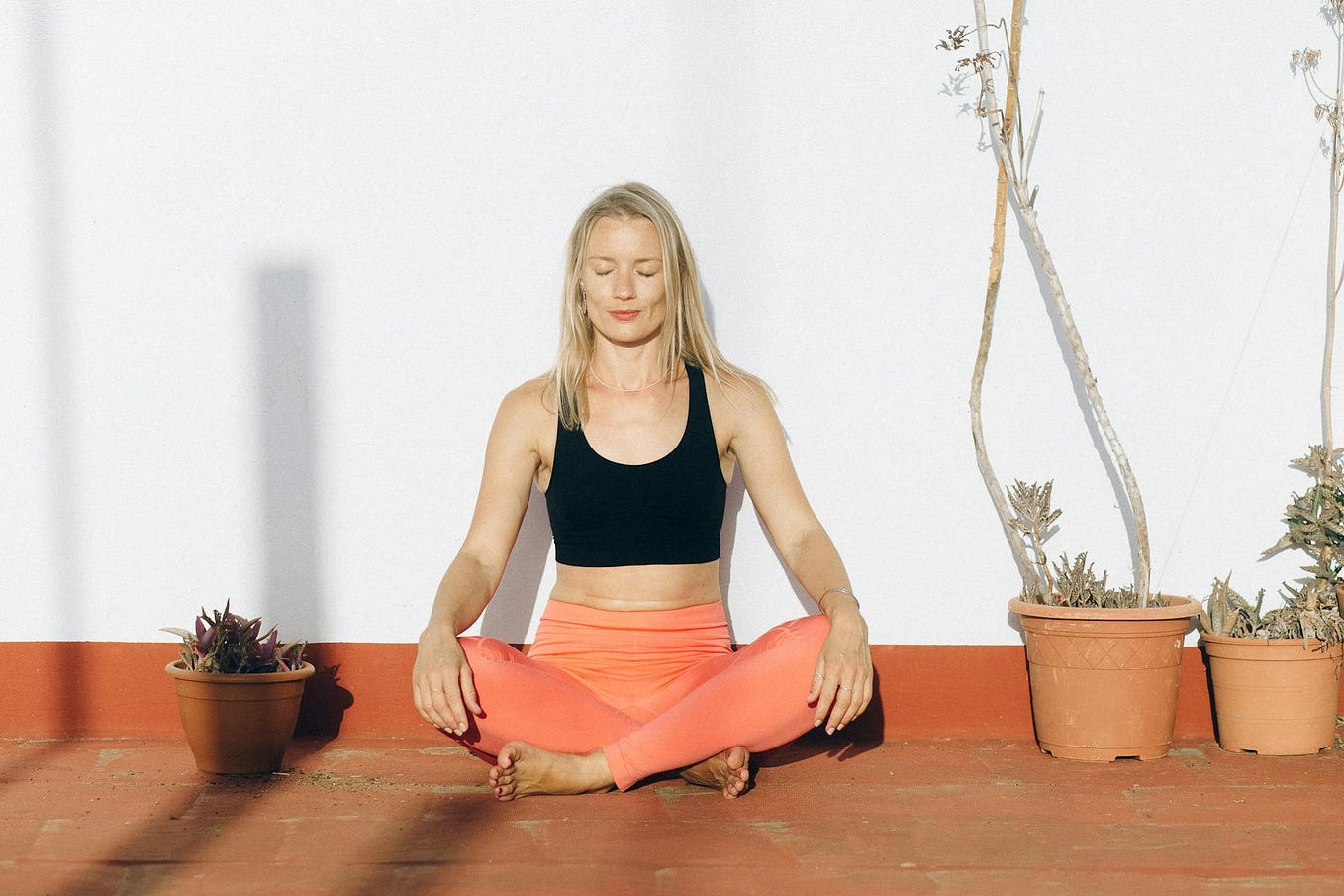 Woman in coral exercise pants sitting with her legs crossed, hands on her knees, and eyes closed.