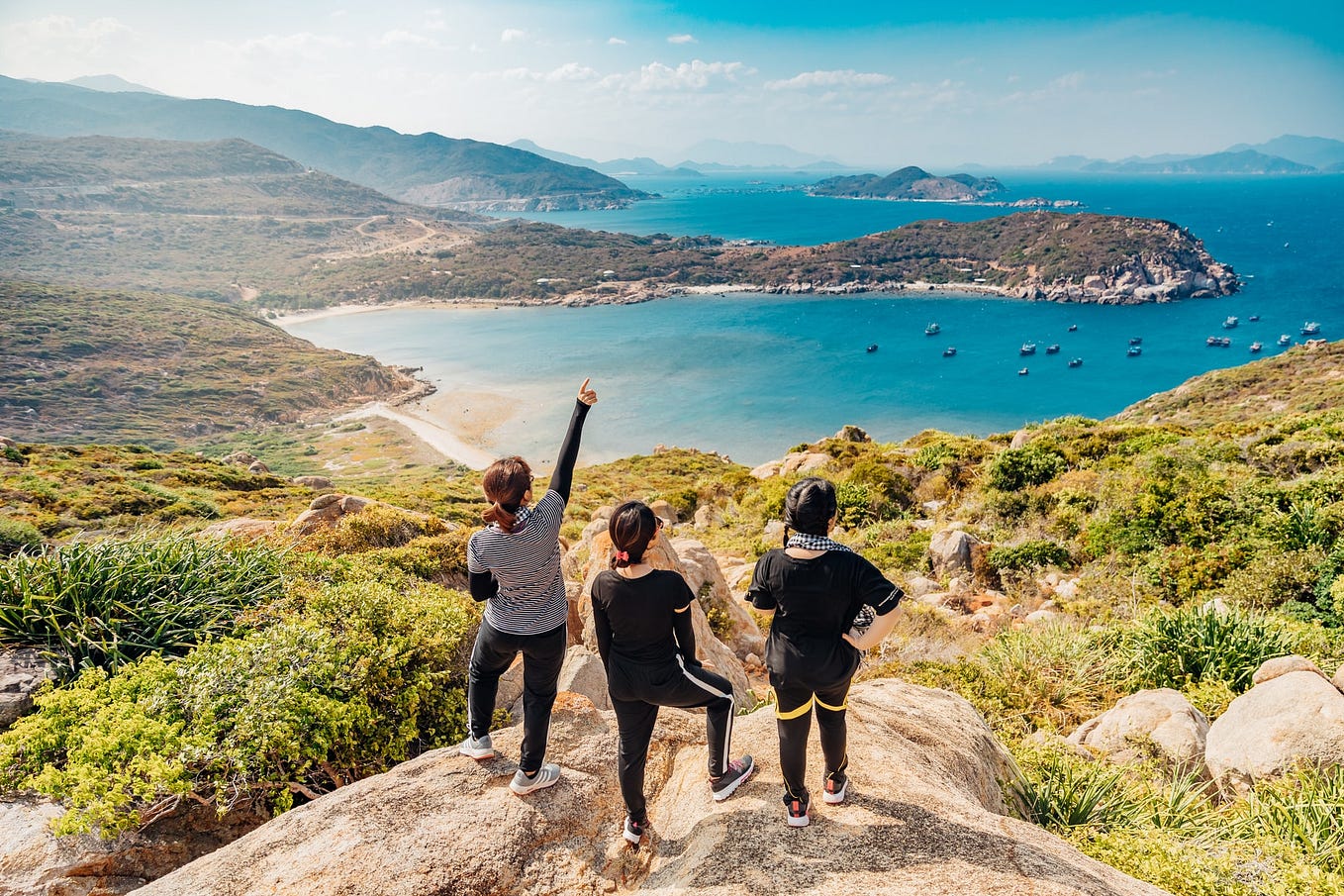 Three women on a rocky hilltop looking out over a sunlit ocean bay