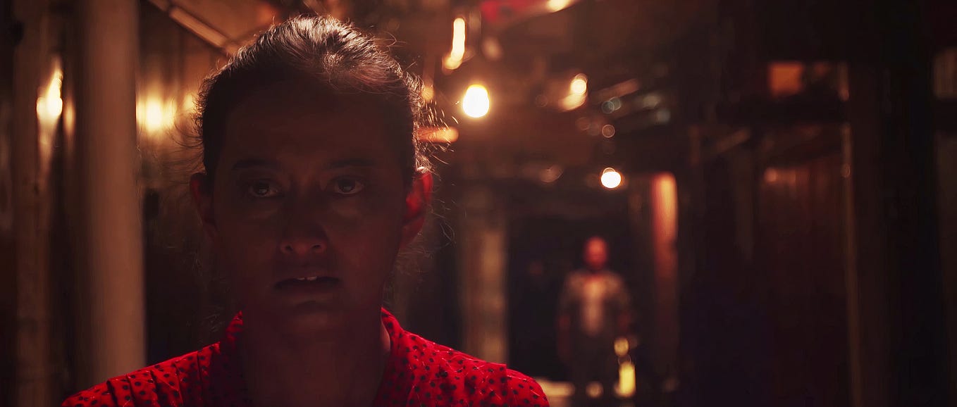 Film still from RONG of protagonist played by Maryam Supraba, wearing a red dress, walking toward the camera through a dark alleyway, followed by a man at a distance, just out of focus.