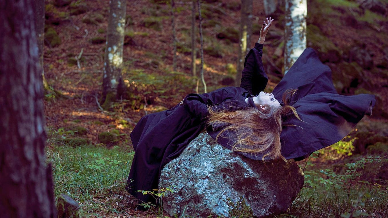 Photo of a woman dressed as a witch laying on a rock in a forest.