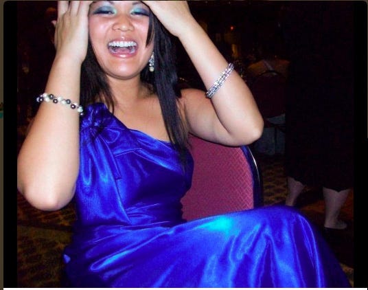Ronni Percy, mid-laugh while fixing her hair in a one-shoulder royal blue gown. A diamond bangle adorns on the left forearm while a multi-colored pearl bracelet sits on the right forearm.