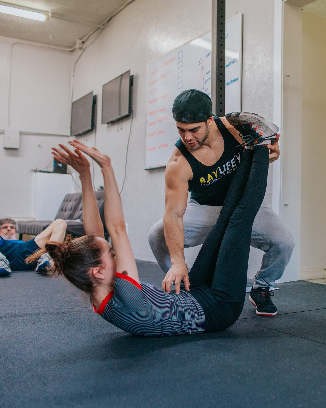 A male trainer at a gym helps a young woman stretch, an older man in the background looks on.