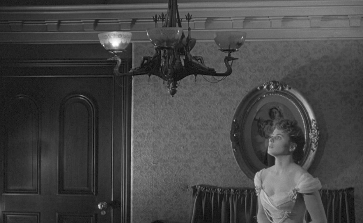 A black and white still from the 1944 film Gaslight. Paula, played by Ingrid Bergman, stares up with a haunted expression at a flickering gaslight chandelier in her home. Just one of its light is on.