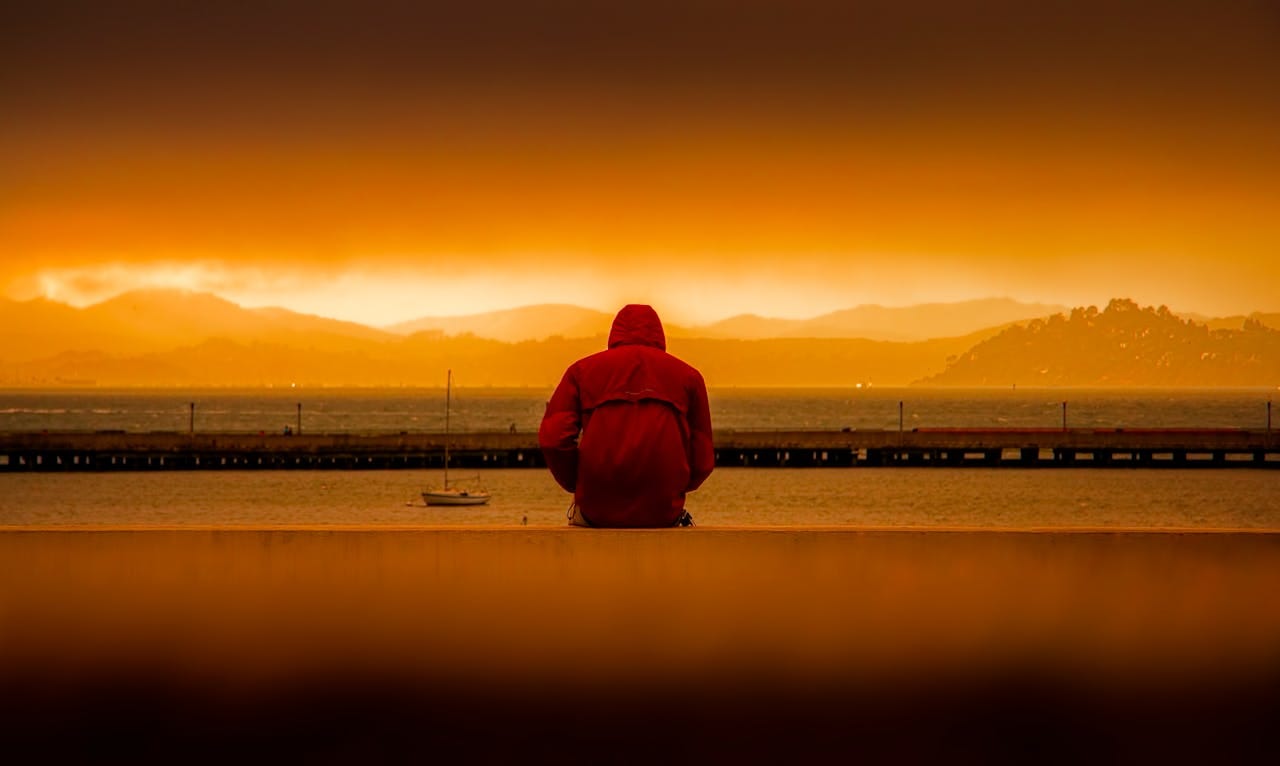 A person in a red jacket looks out at an orange sunset over water.