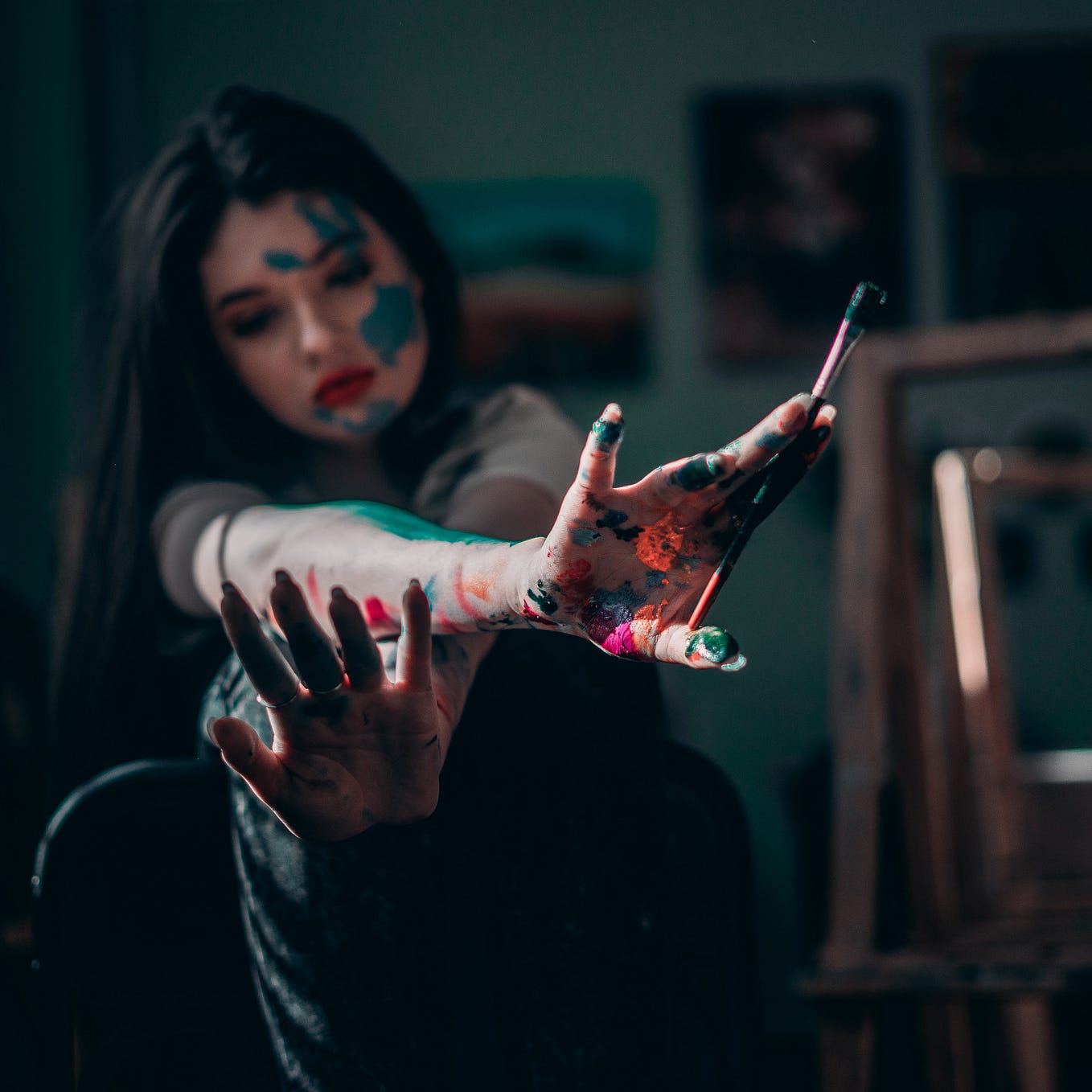 A woman covered in paint and holding a paint brush; her face is blurred in the background, with her hands in the foreground of the photo. She appears to be studying them.