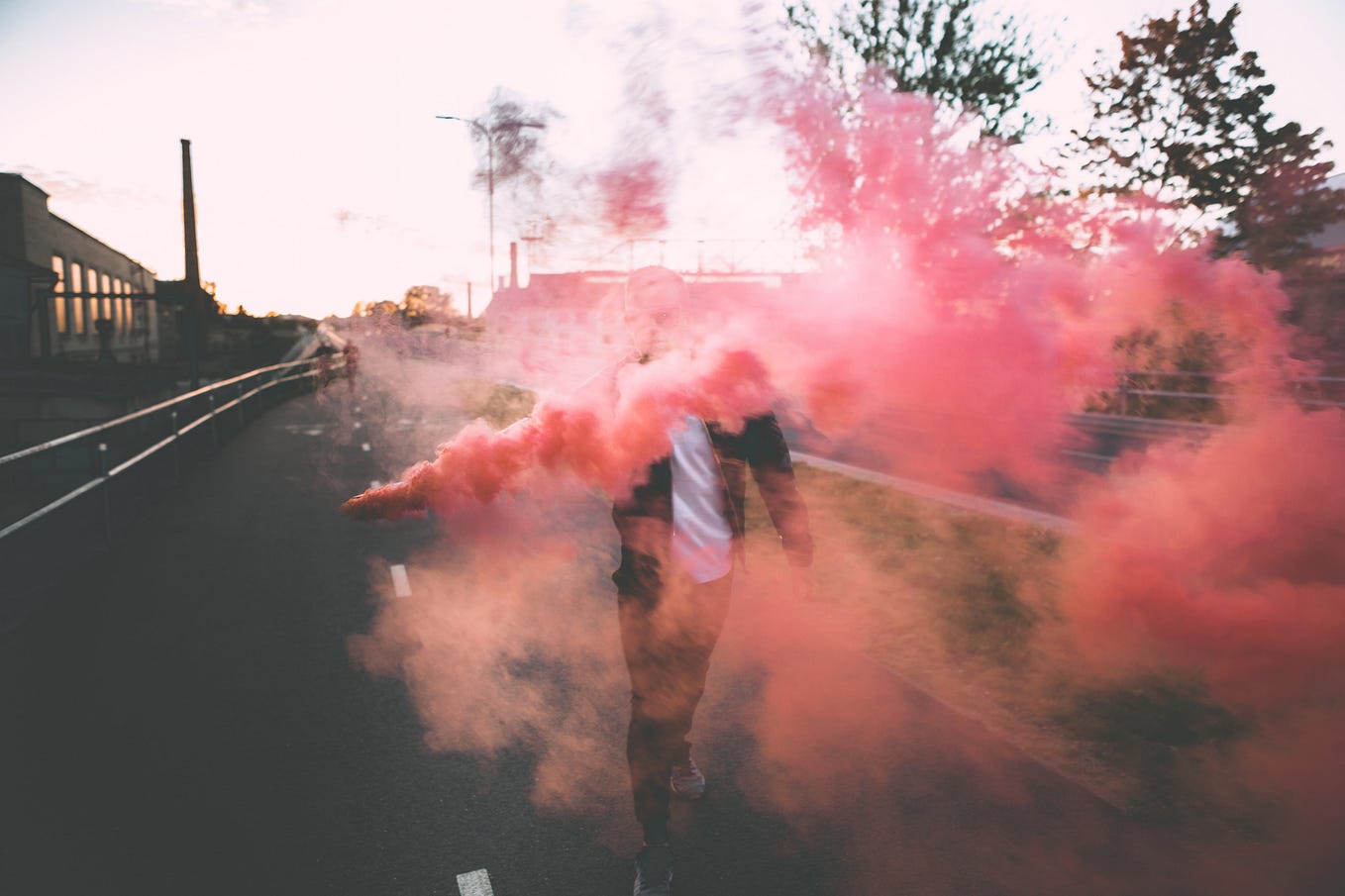 Person in a leather jacket standing in the street with colorful red smoke bomb obscuring face.