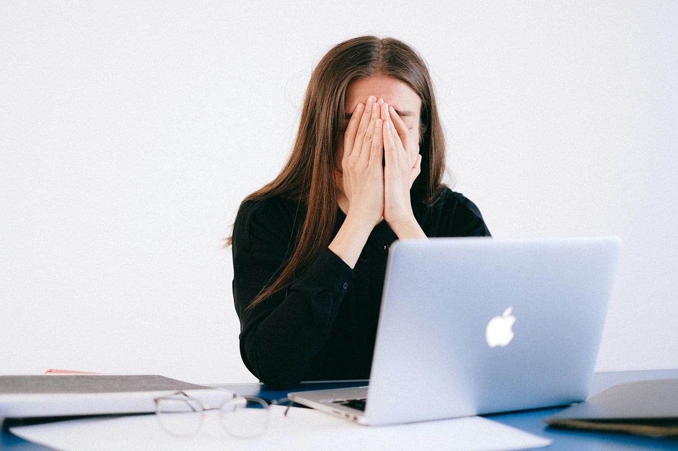 Dark-haired white woman sits in front of Macbook, eyeglasses on the desk, hands pressed over her face, looking stressed or exhausted