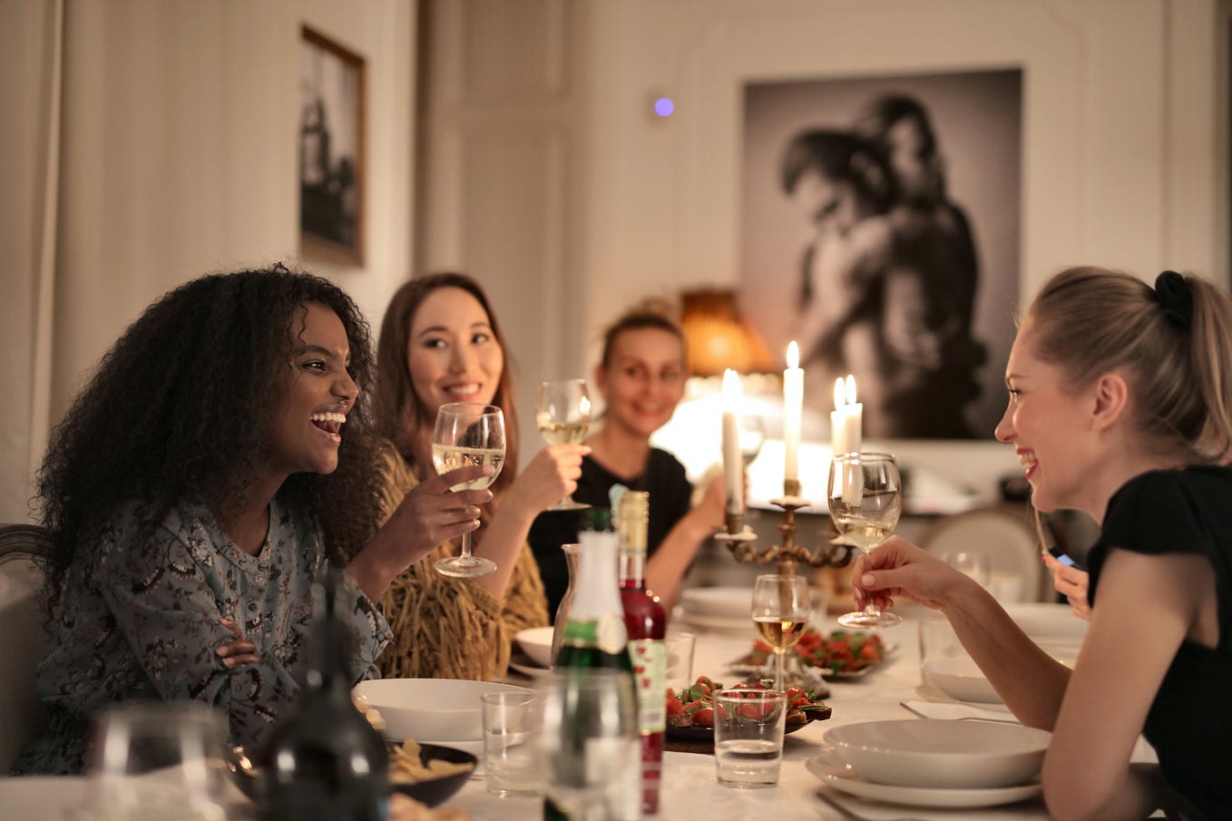 Women Holding Wine Glasses and laughing. They’re at a candlelit dinner party.