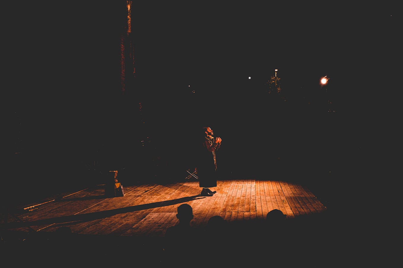 An image of an actor in the centre of a spotlighted stage. The floor is wooden, and the rest of the scene is very dark apart from a few heads of an audience in the foreground.