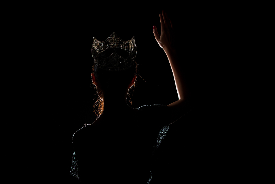 The back of a young beauty pageant contestant standing in the shadowed light wearing the winning crown as she waves to the crowd before her