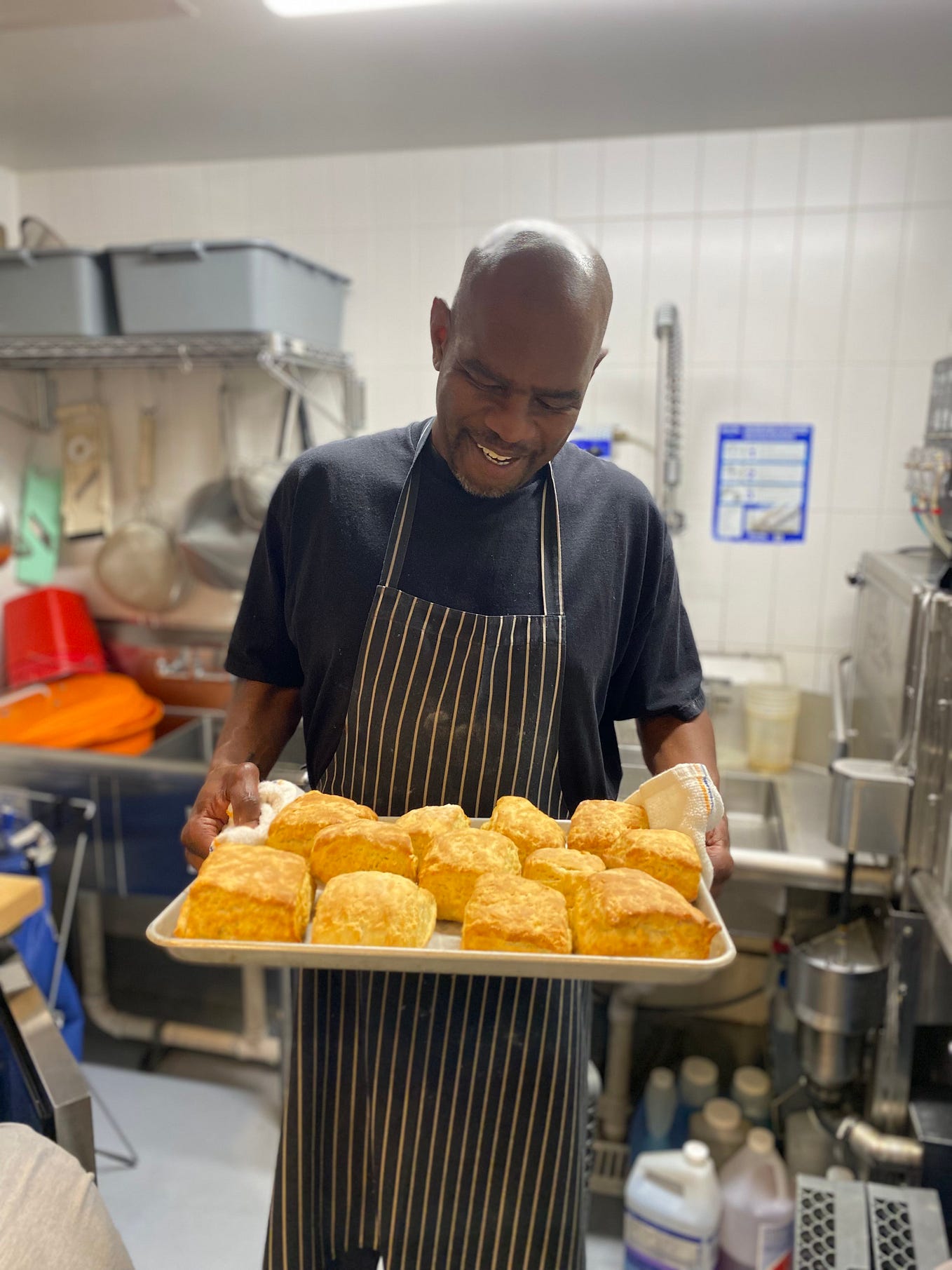 David, a black man, stands smiling down at a baking sheet filled with biscuits. He is wearing a black t-shirt and a black and white striped apron in an industrial kitchen.