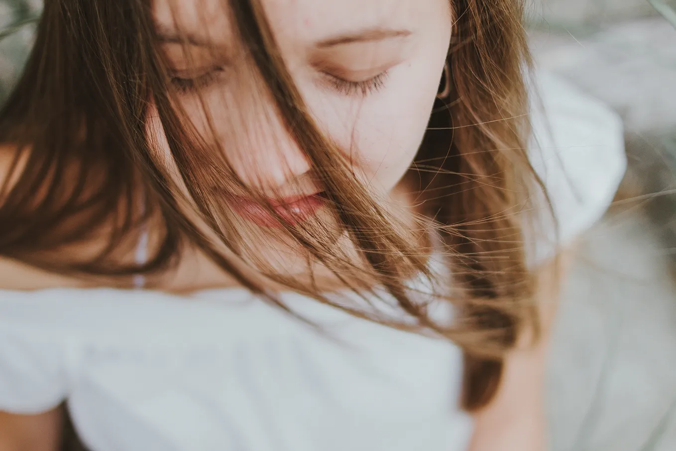 A young white woman, as seen in close up. She has brunette hair extending just below her shoulders. She wears a white dress and her eyes are closed as her hair falls across her face.